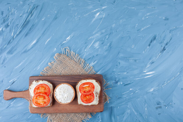 番茄西红柿片放在奶酪面包上 一碗面粉放在切菜板上 粗麻布餐巾纸放在蓝色布上开胃菜美味切片