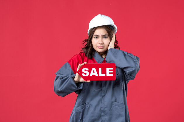 肖像正面近距离观看穿着制服 戴着安全帽的紧张女工人在隔离的红墙上展示销售图标吸引硬穿着
