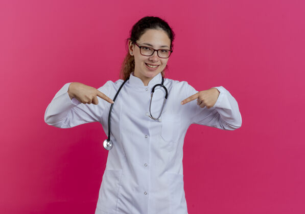 眼镜面带微笑的年轻女医生穿着医用长袍 戴着听诊器和眼镜 指着隔离在粉红色墙上的自己医疗指着听诊器