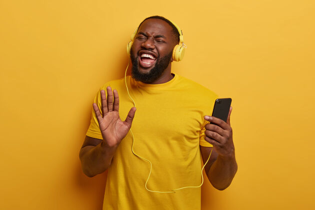 设备精力充沛的美国黑人男子拉着手掌对着相机 用智能手机和耳机收听广播或播放列表中的音频曲目 用最喜欢的歌曲提升心情耳机男人积极