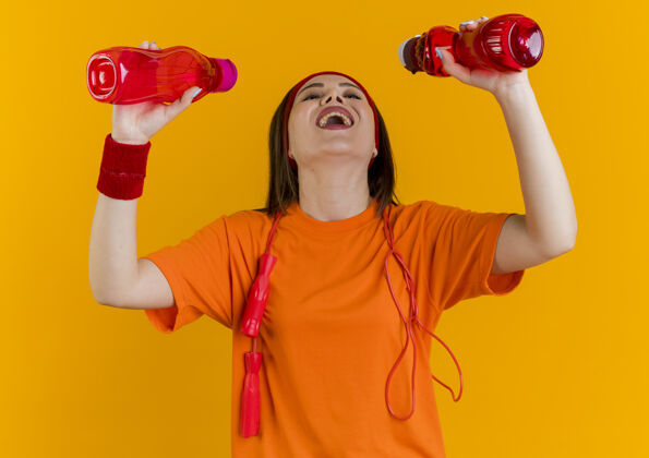 头带快乐的年轻运动女性戴着头带和腕带 脖子上系着跳绳 看着准备喝水的瓶子欢乐看运动