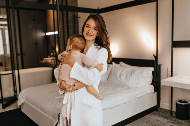 女儿穿着浴衣的年轻母亲抱着尿布里的婴儿 对着白色的床微笑着看着摄像机年轻浴袍妈妈