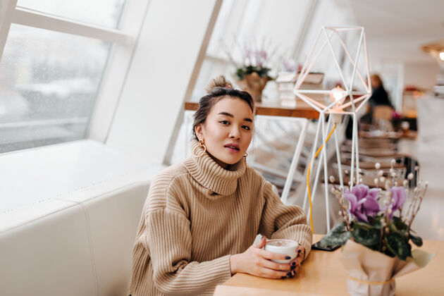 微笑穿着米色毛衣拿着一杯咖啡的时尚亚洲女人的肖像咖啡馆皮肤棕色眼睛
