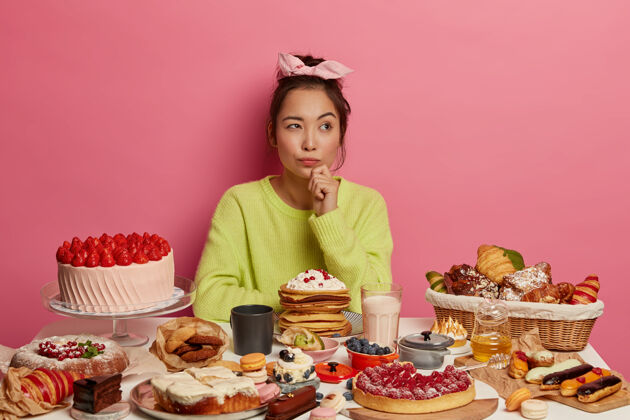 沉思沉思的韩国女士喜欢美味的小吃 吃着美味的糕点 蛋糕和薄饼 想着如何戒掉甜食瘾 抱着下巴 摆着手工糖果在节日餐桌上桌子思考头带
