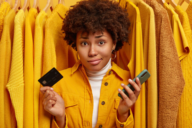 卷发不高兴的不知情的女人卷发 无法支付所有的钱买衣服 拿着塑料卡和现代手机 对着衣架上的普通黄色套头衫摆姿势商店销售机架