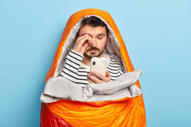 装备疲惫的男性露营者揉眼睛 使用手机 试图在野外连接到INTERNET 在睡袋中摆姿势 拥有露营所需的所有必要设备科技胡子冒险