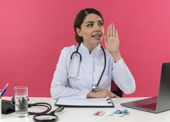 女高兴的年轻女医生穿着医用长袍和听诊器坐在办公桌旁 手持医疗工具和笔记本电脑 一边看着一边把手举在空中笔记本电脑办公桌医生