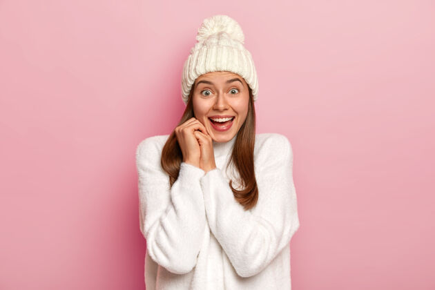 微笑快乐的黑发千禧一代女孩对好消息有快乐的反应 笑容灿烂 戴着温暖的冬帽和舒适的白色毛衣 有热情的目光 孤立在粉色的墙上肖像头饰帽子
