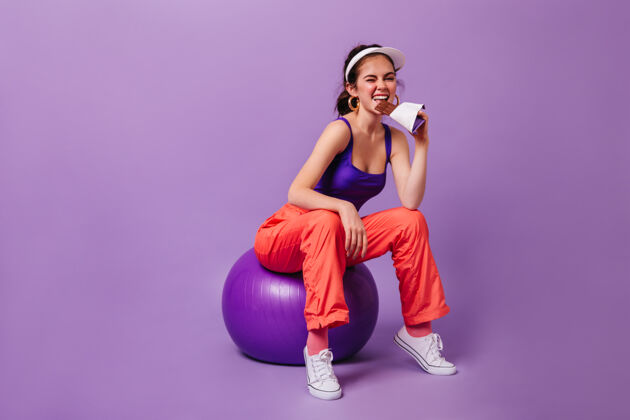 连体衣穿着紫色上衣和红色运动裤的时髦女人坐在紫色墙上的fitball上吃巧克力运动健身球健康