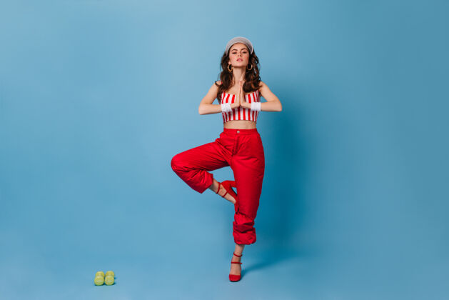 时尚穿着红色裤子和条纹不规则上衣的时髦女士站在蓝色墙壁上的树形姿势和绿色哑铃明亮红色裤子优雅