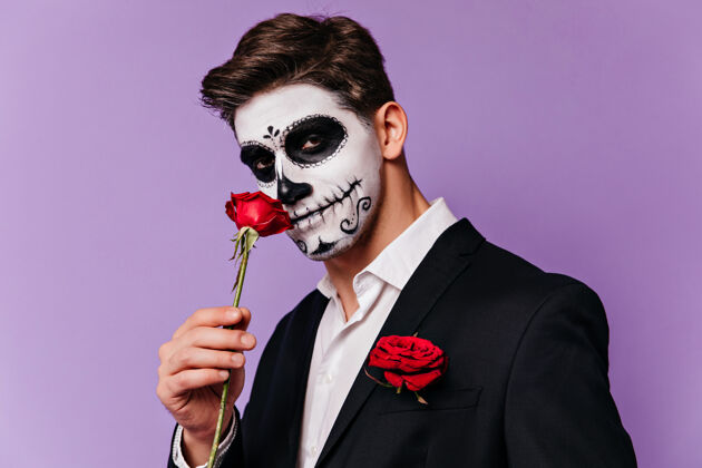 玫瑰穿着燕尾服的黑发男人在万圣节和罗斯合影漂亮的男模 带着墨西哥恐怖的化妆品站在紫色的背景上装饰品幽灵节日