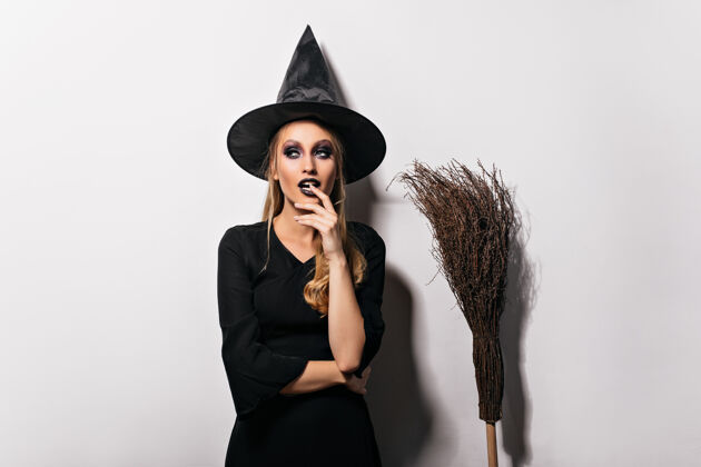 庆典沉思的女巫师在白墙上摆姿势戴着黑帽子的性感小巫婆站在扫帚旁边拼写魔鬼金发