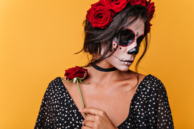 幻想迷茫的浪漫女孩 头骨造型的面部艺术 锁骨上抱着玫瑰 橘色墙壁上黑发女郎摆着黑色上衣花人物民间传说