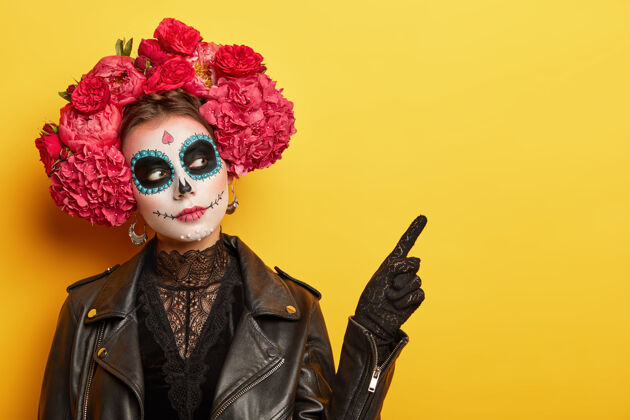 油漆严肃的年轻女士戴着死亡面具头盖骨化妆 花环 点在黄色墙壁上的自由空间幽灵花环指标