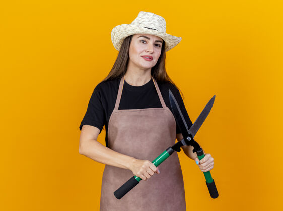 制服高兴美丽的园丁女孩穿着制服和园艺帽子拿着剪子孤立在橙色手握帽子剪子
