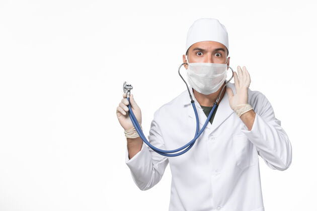 专业正面图：男医生穿着医疗服 戴着口罩 由于冠状病毒-使用听诊器对白色墙壁冠状病毒-大流行疾病冠状病毒药品冠状病毒预防
