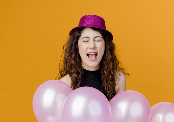 尖叫年轻漂亮的女人 卷发 戴着节日礼帽 手里拿着一堆气球 疯狂地尖叫着生日派对的概念站在橘色的墙上疯狂一群站着