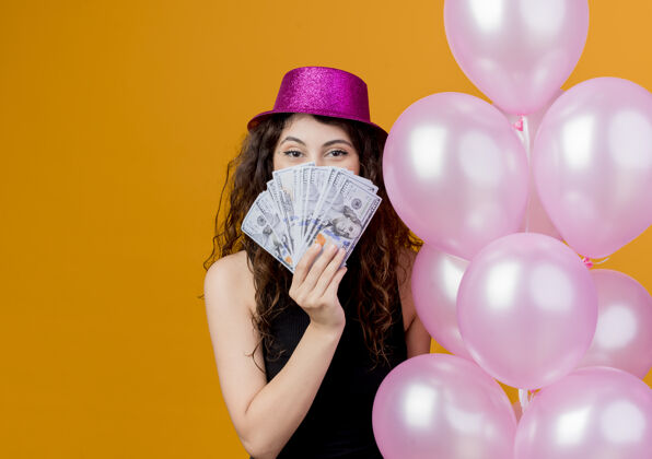 束一位年轻漂亮的女士 一头卷发 戴着节日礼帽 手里拿着一堆气球 站在橙色的墙上 展示着现金快乐和兴奋的生日派对理念年轻女人空气