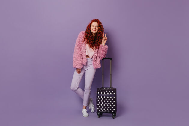 圆点一头卷曲的红发 身着明媚的春装 在紫色的空间里展示着和平的标志和手提箱的姿势魅力上衣手提包
