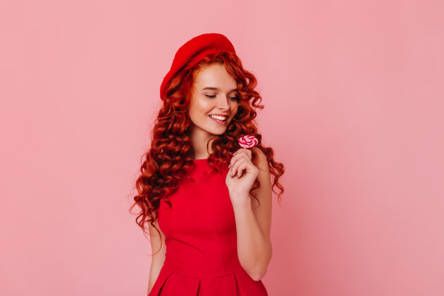 奢华可爱的年轻女子穿着红色连衣裙 戴着毡帽 在粉红色的空间里摆着棒棒糖的姿势头发少女自信