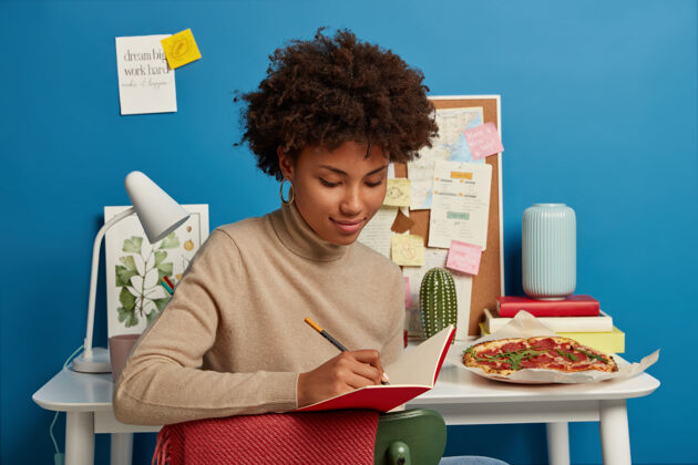 比萨饼漂亮的女性留着非洲发型 在记事本上记笔记 写下自己的想法 坐在白色桌面旁边的椅子上 带着工作 学习 教育的必备物品女学生书桌卷发