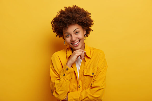 微笑自然美的可爱女人肖像 非洲式发型 穿着亮黄色夹克 戴着大耳环 摸着下巴 表情悠闲 在室内摄影棚摆姿势非洲高兴下巴