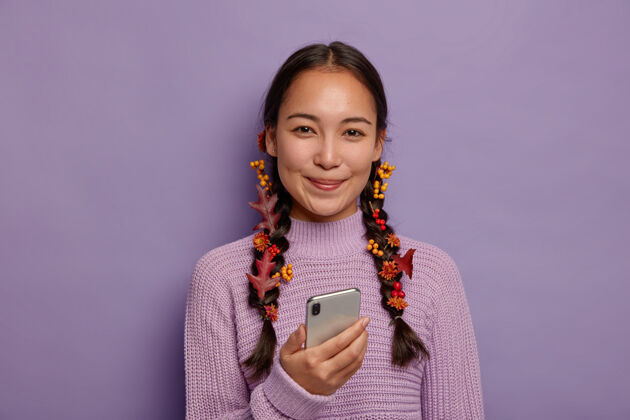 微笑美丽的季节概念迷人的亚洲女士拥有自然的美丽 深色的头发和秋天的落叶梳成两个辫子 享受秋天的舒适 在休闲时间使用现代智能手机女孩编织温柔