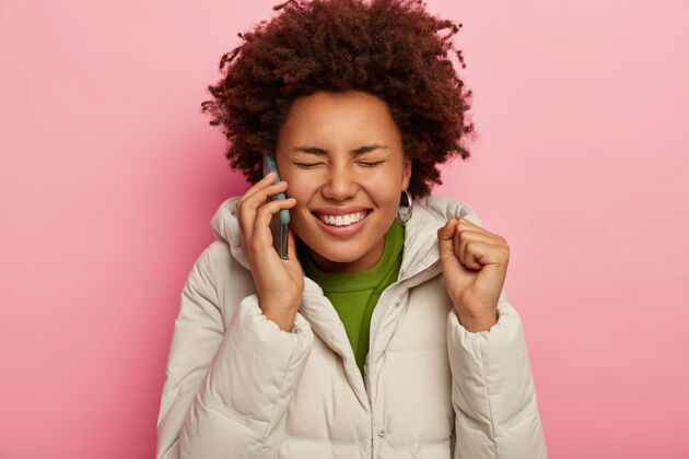 黑色漂亮的卷发女人通过现代智能手机打电话给朋友 举起紧握的拳头 笑容灿烂 身穿带兜帽的白色夹克 粉色背景下的模特说话电话粉色