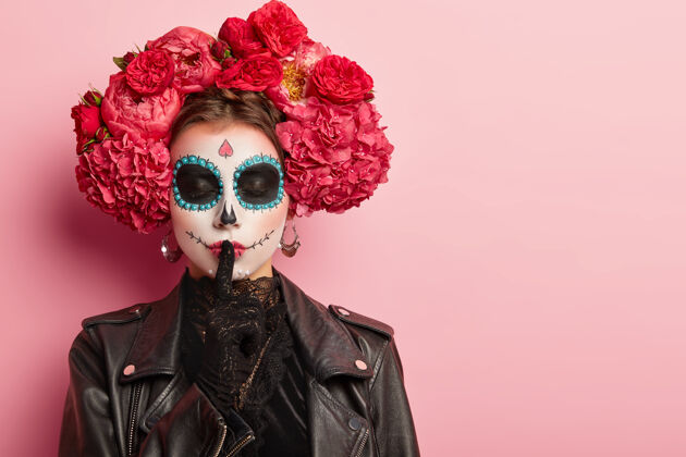 墨西哥人有创意化妆的女性水平镜头 身着黑色服装 展示沉默的手势 闭上眼睛 对着粉色墙壁摆姿势空白空间粉色秘密