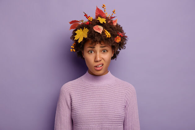 忧郁可爱的不满的非裔美国女士微笑着面对 钱包嘴唇 有悲伤的脸表情 穿着黄色的叶子和浆果在头发针织毛衣孤立的紫色美国秋天紫色