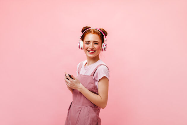 女性带着发髻的年轻女士惊讶地看着手机屏幕穿着裙子和白色t恤的女士正在用粉色背景的耳机听音乐成人肖像优雅
