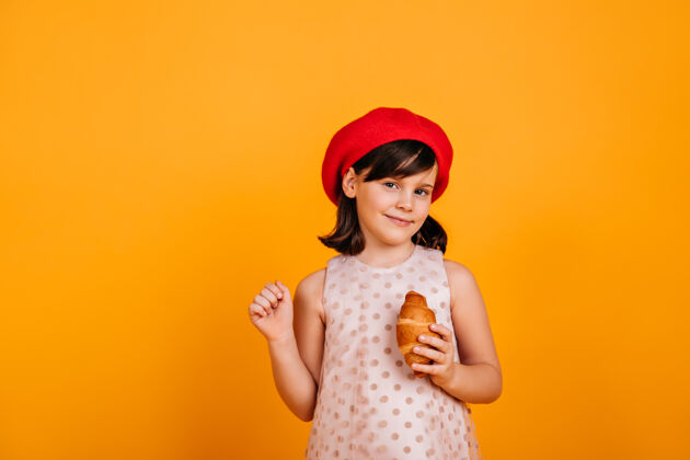 乐趣好奇的黑发小孩在黄色的墙上摆姿势吃羊角面包的未成年女孩羊角面包可爱模型