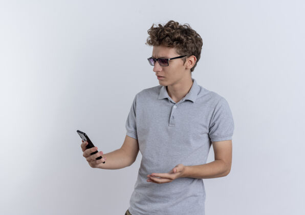 站穿着灰色马球衫的年轻人站在白色的墙上看着他的智能手机屏幕 看起来很困惑移动马球屏幕