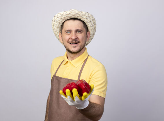 微笑年轻的园丁穿着连体衣 戴着帽子 戴着工作手套 脸上露出红甜椒的笑脸手套红色表演