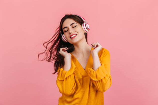 享受活泼的女人带着雪白的笑容在粉红色的墙上跳舞穿着橙色上衣的模特在听音乐耳机女孩裤子