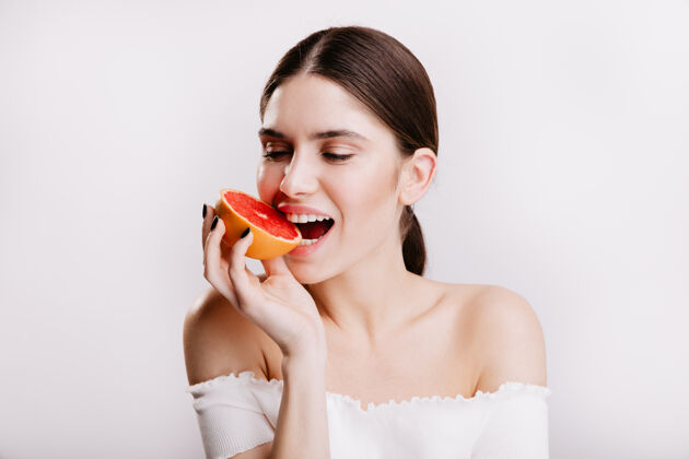 眉毛皮肤健康的黑发女孩咬着多汁的红葡萄柚隔离墙上的女人心情很好的特写照片看起来柑橘完美