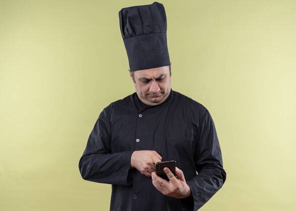 帽子男厨师身穿黑色制服 头戴厨师帽 手持智能手机向站在绿色背景下严肃表情的人发短信绿色烹饪认真