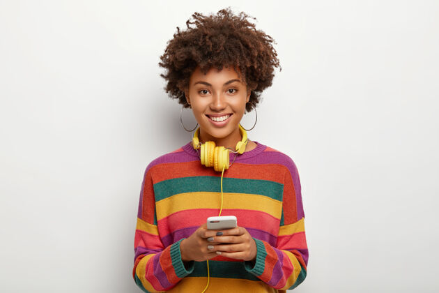 欢呼正高兴的非裔美国妇女身着条纹彩色毛衣 手持连接耳机的现代手机 上网冲浪高兴互联网耳环