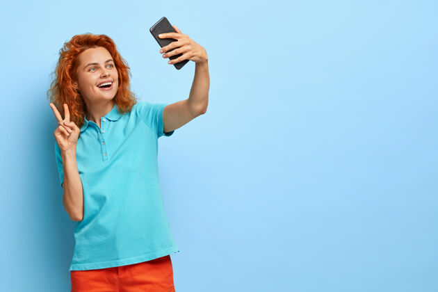 看姜黄色快乐的卷发女人 做和平手势 在智能手机上自拍 玩手机 穿t恤自己穿自拍