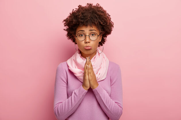 人黑皮肤 非洲发型的年轻女子照片 表情可怜 双手合十 要求再给一次机会 相信更好 戴圆眼镜 紫色套头衫祈祷室内眼镜