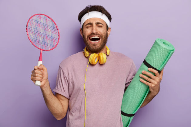 训练情绪化的运动型男子用健身垫和网球拍训练 笑得表情喜出望外 黑发 穿着休闲服 训练时听音乐教练微笑紫色