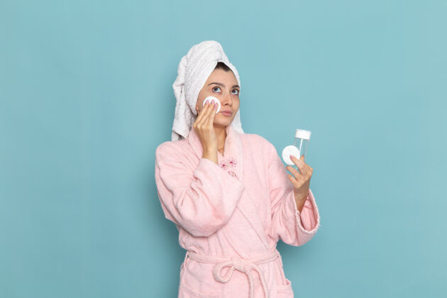 化妆品正面图身着粉色浴袍的年轻女性在蓝色墙壁上洗脸化妆清洁美丽净水自我护理霜淋浴前面年轻人奶油