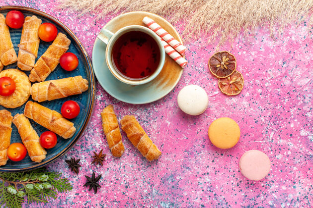 生的顶视图甜甜可口的百吉饼在托盘内与新鲜的酸李子麦卡龙和一杯茶在淡粉色的桌子上甜点饼干饼干