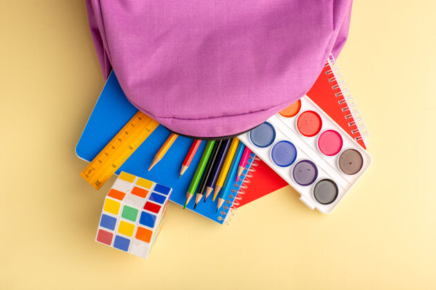 油漆顶视图彩色铅笔与抄写本油漆和浅黄色书桌上的紫色包学校毛毡笔铅笔本记事本容器抄写本学校
