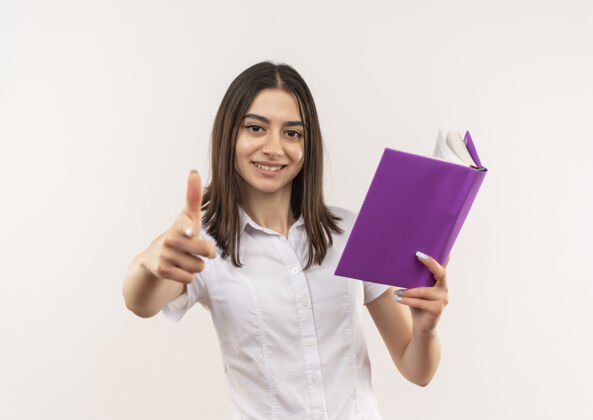 手势穿着白衬衫的年轻女学生站在白墙上 手里拿着一本打开的书 微笑着竖起大拇指人女站着