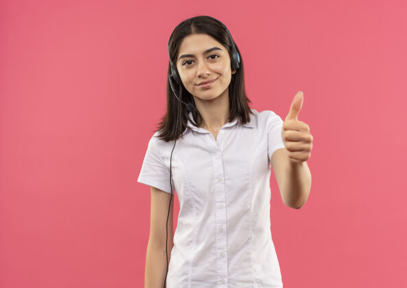 手势穿着白衬衫 戴着耳机的年轻女孩 微笑着站在粉色的墙上 竖起大拇指望着前面站微笑公民