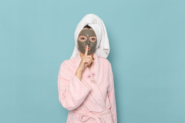 脸正面图身着粉色浴袍的年轻女性 脸上戴着面具 蓝色墙壁上的“沉默”标志淋浴清洁美容自理霜沉默展示前面