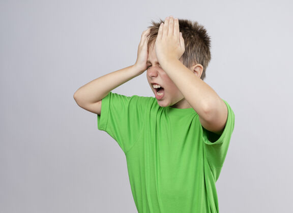 感觉生病的小男孩穿着绿色t恤 身体不适 头痛得厉害 站在白墙上碰头头疾病不适