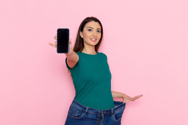 运动正面图身着绿色t恤的年轻女性手持智能手机在浅粉色墙壁上腰部运动锻炼美丽苗条女性运动减肥腰部运动