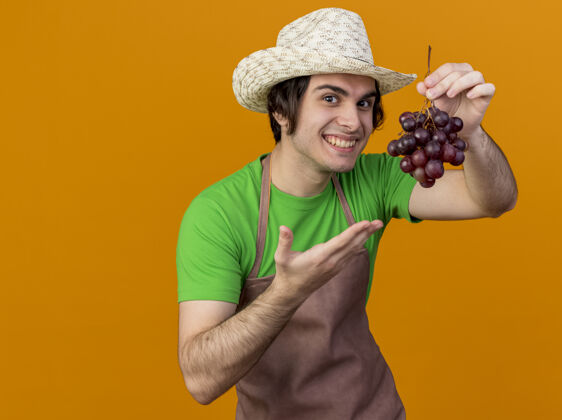 介绍年轻的园丁 围裙戴帽子 手里拿着一束葡萄 微笑着站在橘色的墙上橘子葡萄展示